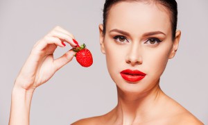 Sweet temptation. Beautiful young shirtless woman holding strawberry in her hand while standing against grey background
